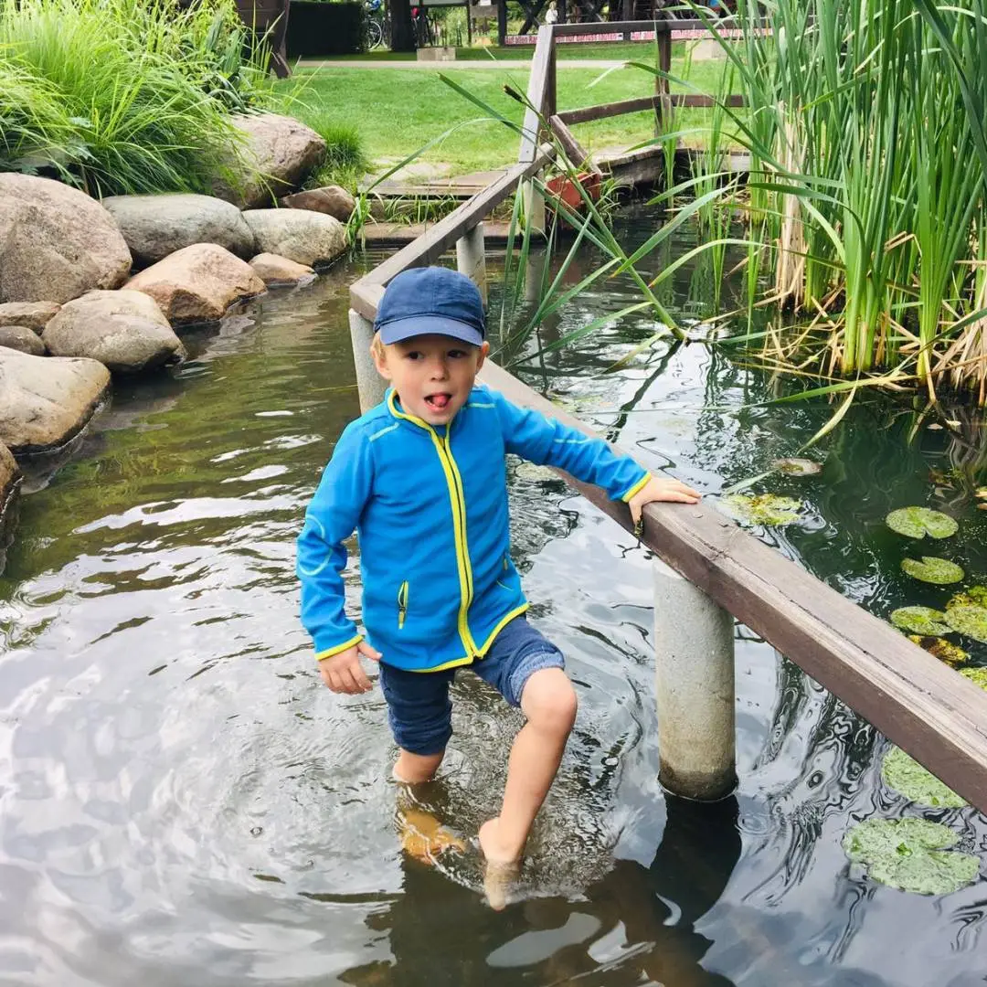 Kind mit blauer Mütze und blauer Jacke watet durch das flache Wasser eines Naturteichs