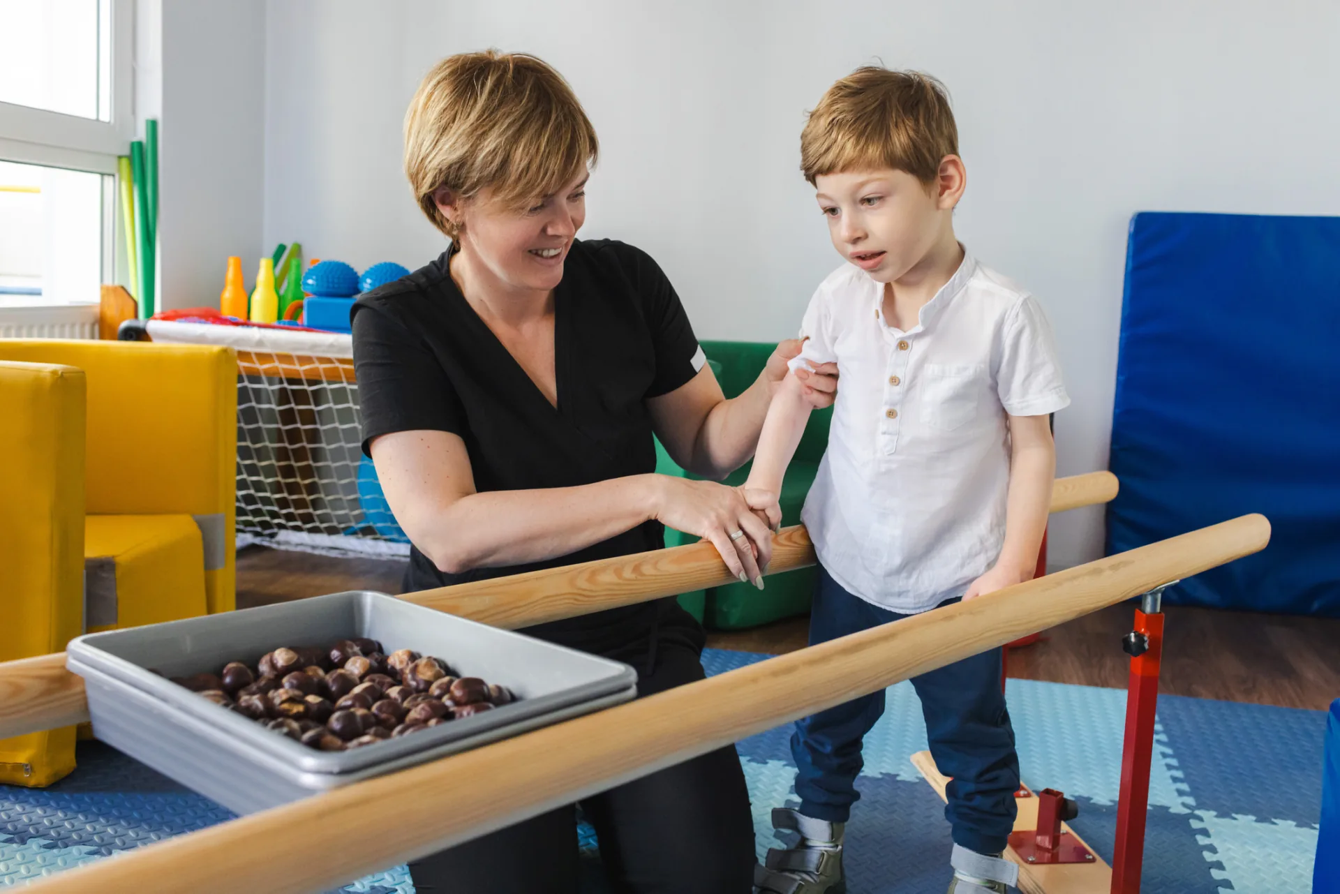 Therapeutin führt den Arm eines Jungen an einer Holzstange, davor eine Wanne mit Kastanien im bunten Therapieraum