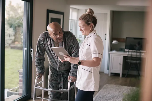 Pflegekraft mit Tablet im Gespräch mit einem Patienten am Rollator in dessen Wohnung