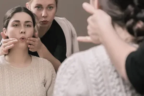 Therapeutin berührt mit den Fingern die Wangen einer jungen Frau, die die Lippen spitzt; im Hintergrund zwei weitere Personen
