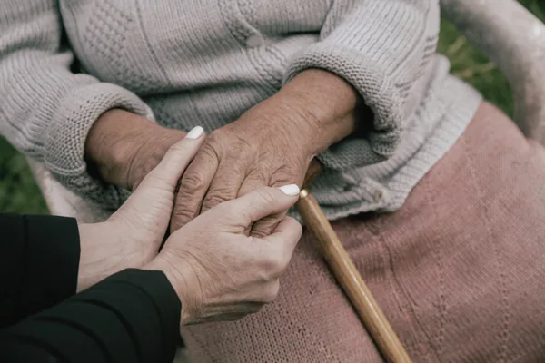 Hände einer Therapeutin stützen die Hände einer älteren Patientin, die einen Holzstab hält